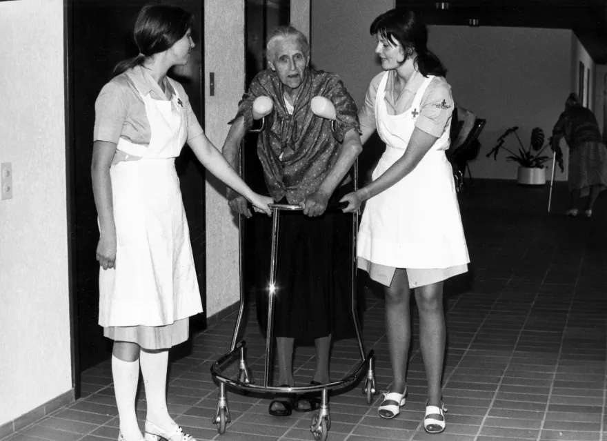 Historisches Bild aus dem Archiv: Zwei Pflegehelferinnen SRK stützen eine alte Frau beim Gehen.